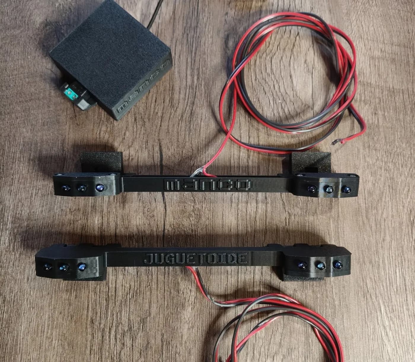 Dos barras IR negras, una con el relieve «manco» y otra con «Juguetoide», con sus cables y una caja negra, sobre una mesa de madera.