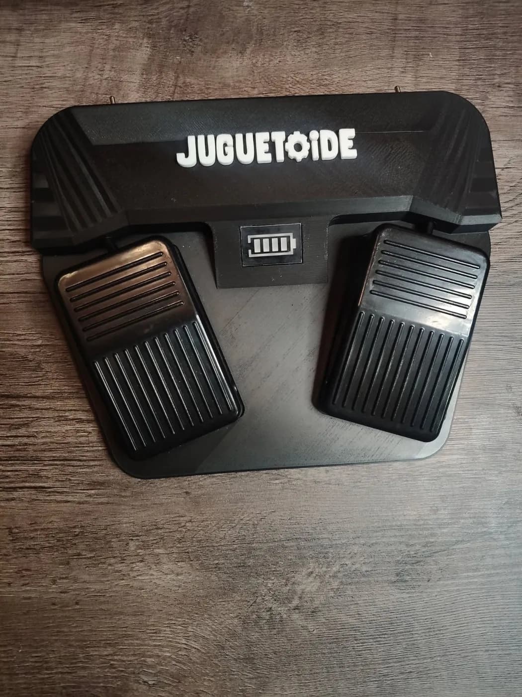 El Cover Pedal visto desde arriba: una base negra con dos pedales, el indicador de batería en el medio y el logo de Juguetoide.