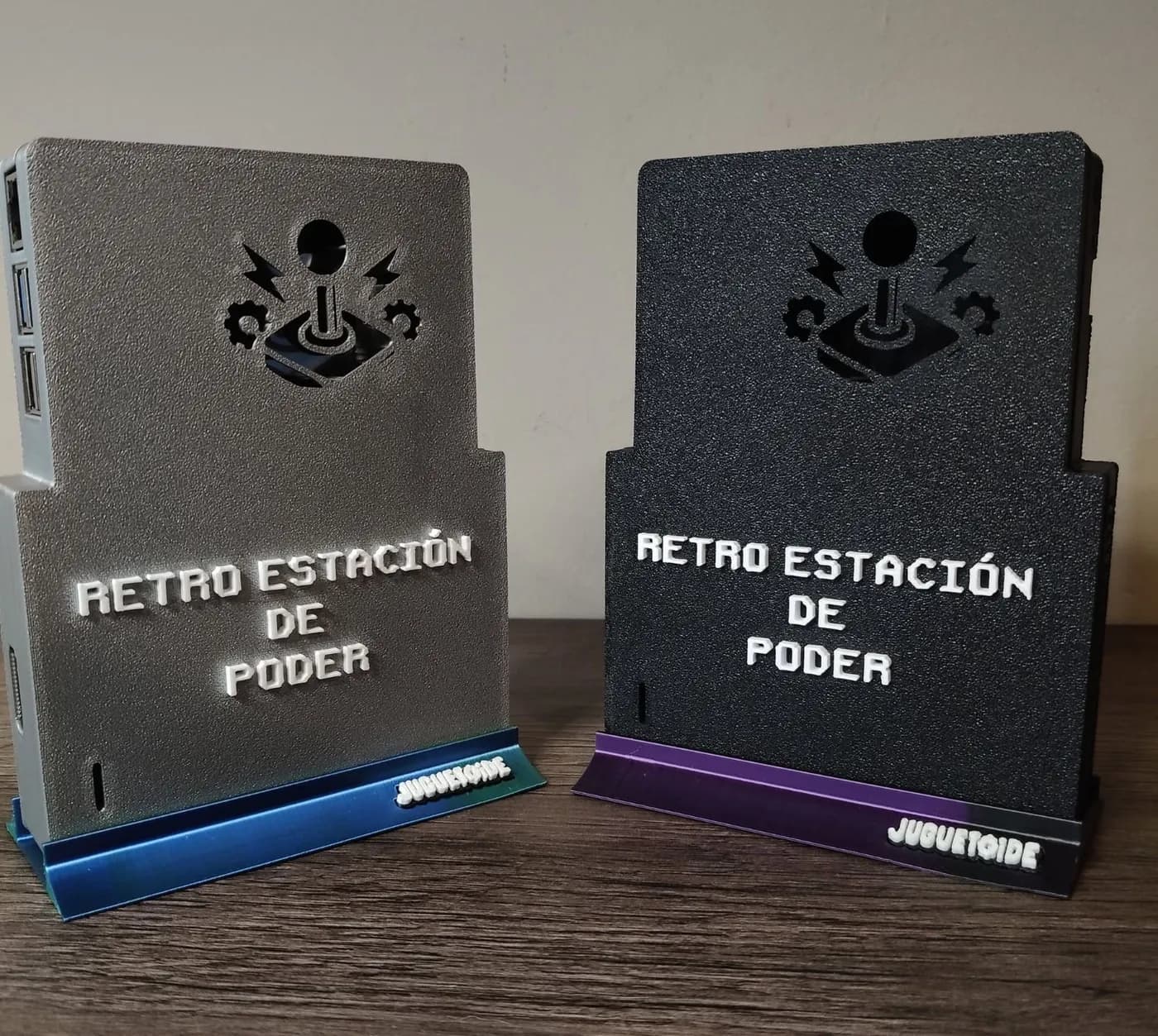 Dos Retro Estación de Poder paradas sobre una mesa, una plateada y una negra, con un joystick calado y el nombre en relieve.