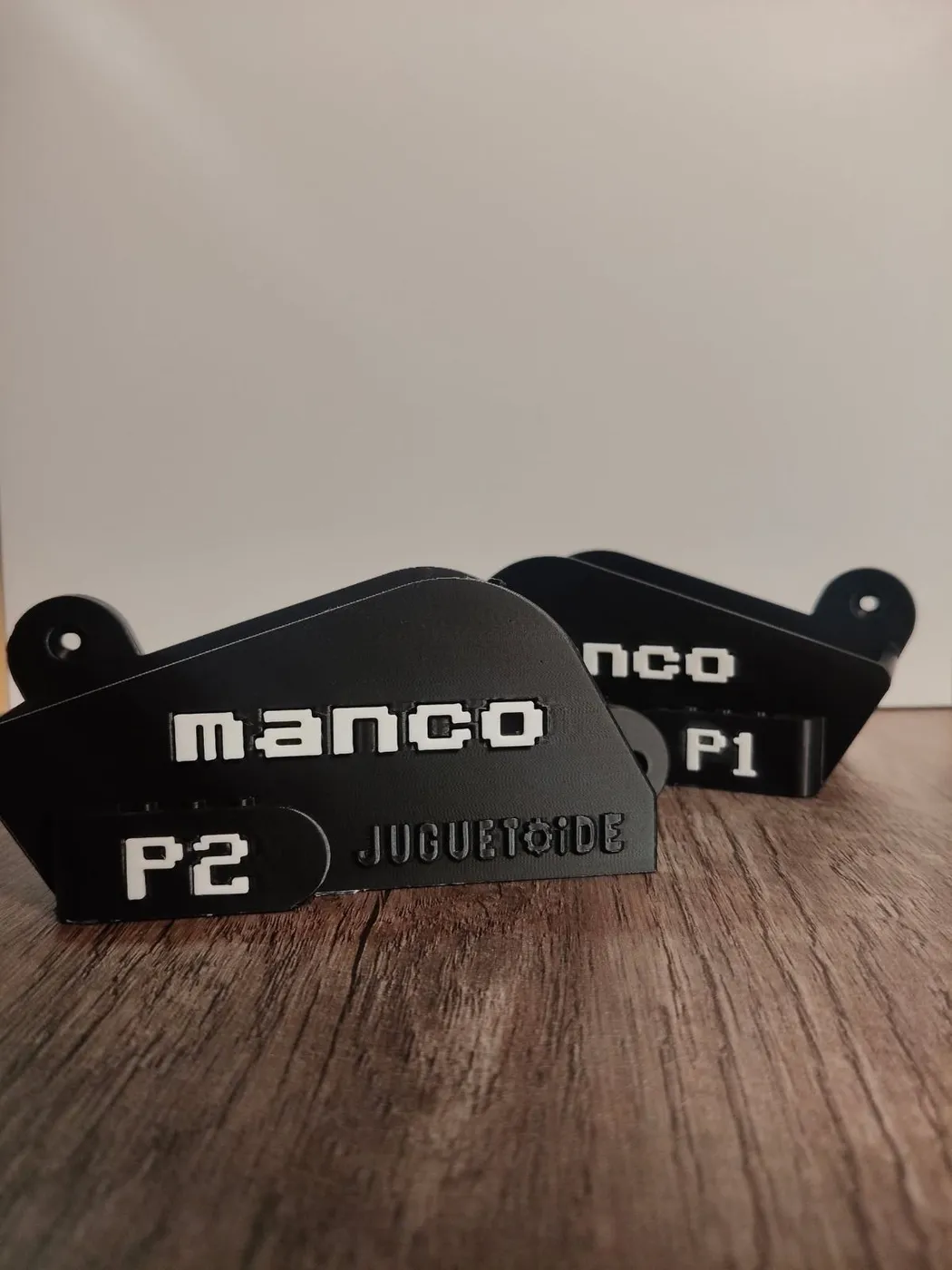Dos soportes negros para la MANCO sobre una mesa de madera, uno rotulado P1 y el otro P2.